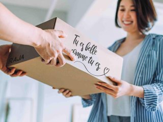 Eine lächelnde Frau erhält ein personalisiertes Paket mit der französischen Aufschrift: Tu es belle comme tu es.